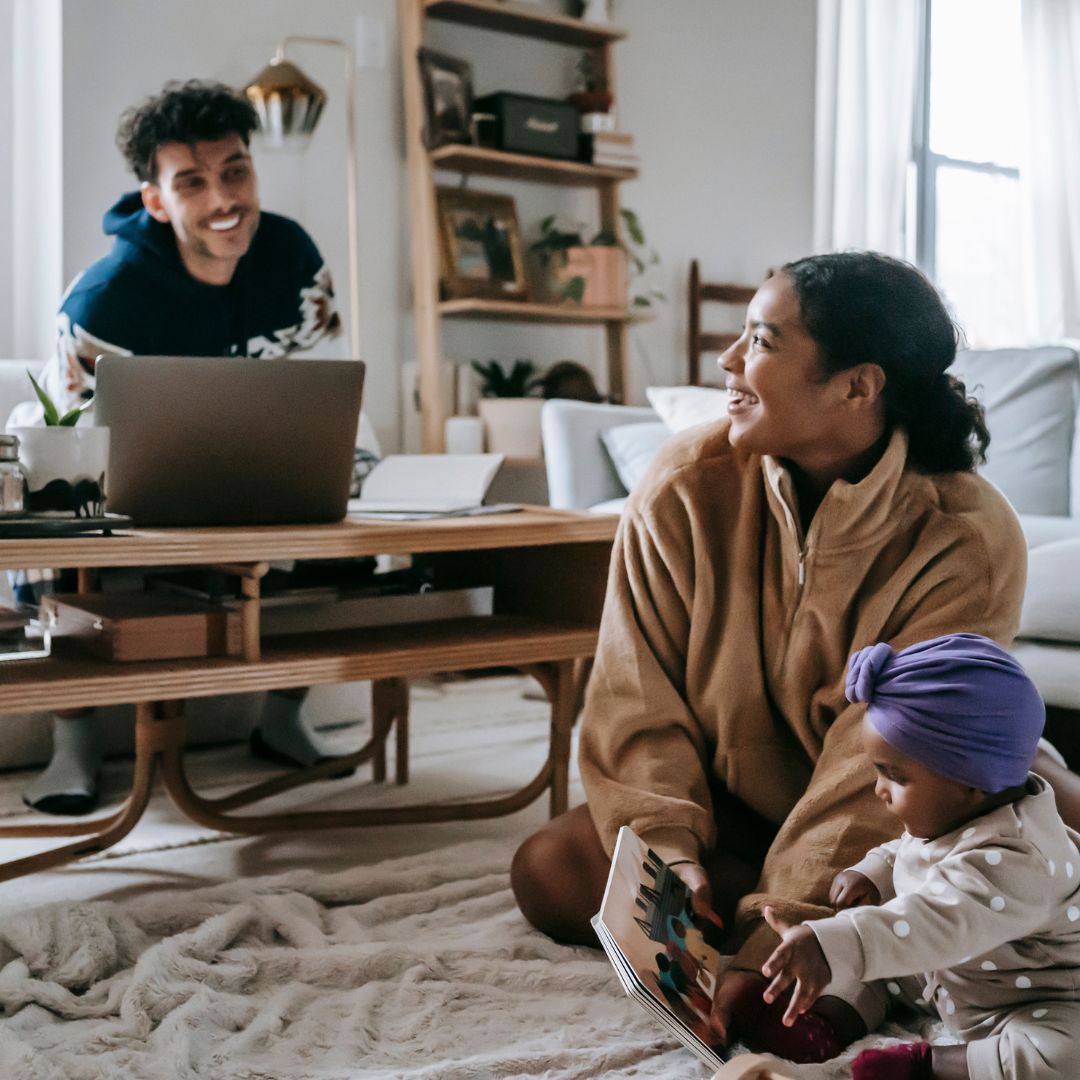 family inside apartment laughing while baby girl is looking at a book